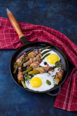 A dish of asparagus, bacon and eggs in a cast-iron skillet on a dark background.