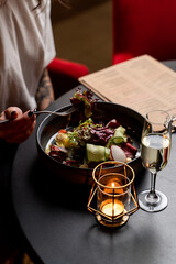 Woman is eating salad with champagne from a black bowl in a restaurant, hands only visible