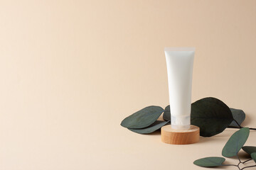 White tube of cosmetic cream with eucalyptus on wooden podium on beige background. Close up, copy space.