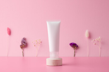 White tube of cosmetic cream with flowers on wooden podium on pink background. Close up.