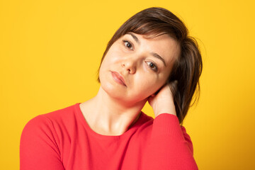 Portrait of good looking woman keeps hand in hair, dressed in casual red jumper isolated over yellow background. Positive human emotions.