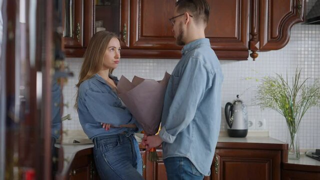 Portrait of young woman with dissatisfied facial expression standing in kitchen as man entering apologizing for mistake with bouquet of flowers. Boyfriend and girlfriend making peace indoors