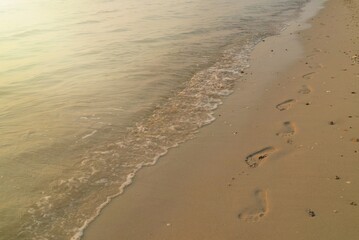 Soft focus of the waves beat at the beach with sand, footprints and reflection of sunlight on water in summer morning. Nature background concept.