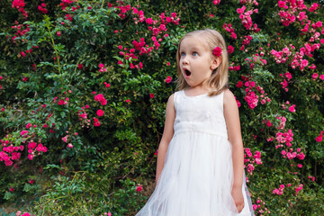 Surprised child with open mouth. Little girl in a white dress on a background of flowering bush