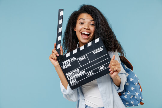 Young cool african american girl teen student in denim clothes backpack holding classic black film making clapperboard isolated on blue background Education in high school university college concept