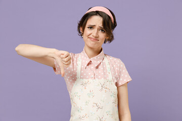 Young disappointed displeased housewife housekeeper chef cook baker woman wear pink apron showing thumb down dislike gesture isolated on pastel violet background studio portrait. Cooking food concept