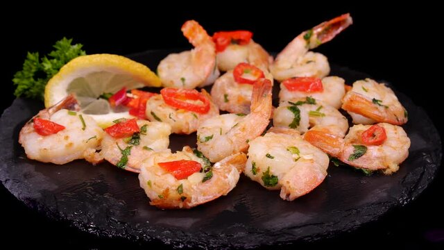 Cooked and grilled shrimps with lemon slice, chilli pepper and parsley rotating on a black plate. Elegant serving of the mediterranean cuisine dish, prawns fried in butter with spices.