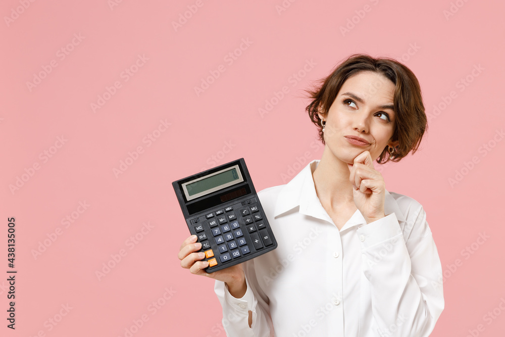 Wall mural Young dreamful successful employee business secretary woman corporate lawyer in classic formal white shirt work in office hold in hands calculator isolated on pastel pink background studio portrait