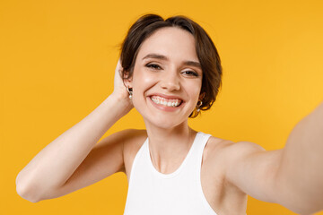 Close up young smiling cheerful happy friendly woman 20s with bob haircut wearing white tank top shirt doing selfie shot on mobile phone hold head isolated on yellow color background studio portrait.