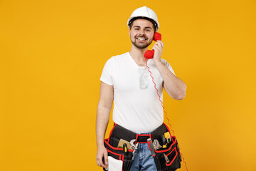 Young happy employee handyman man in protective helmet hardhat speak by handset phone talk isolated on yellow background studio Instruments accessories renovation apartment room Repair home concept