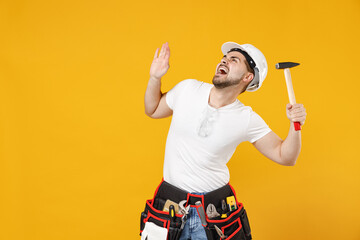 Young scared frightened employee handyman man 20s in protective helmet hardhat glasses hold hammer...