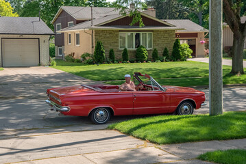 Older man driving his vintage red 1960s convertible in reverse down a residential street.
