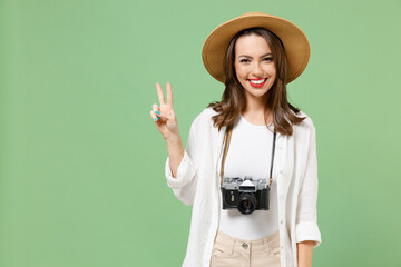 Traveler satisfied tourist woman in casual clothes hat camera on neck show victory v-sign gesture isolated on green background. Passenger travel abroad on weekends getaway. Air flight journey concept.