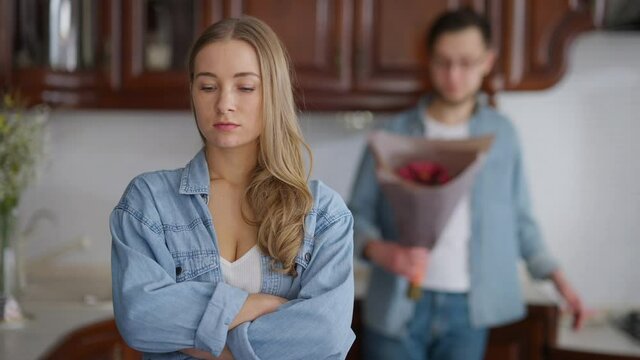Young offended woman looking back at blurred man with bouquet of flowers turning crossing hands sighing. Portrait of angry Caucasian sad girlfriend with boyfriend indoors. Relationship difficulties
