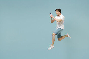 Fototapeta na wymiar Full length side view smiling young man 20s wearing white casual basic t-shirt using mobile cell phone gesture jump high isolated on pastel blue background studio portrait. People lifestyle concept.