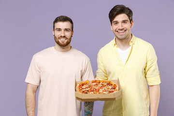 Two smiling happy cool satisifed young men friends together in casual yellow t-shirt hold italian pizza in cardboard flatbox look camera isolated on purple background studio People lifestyle concept.