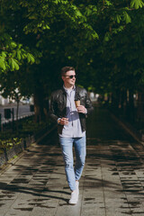 Full length trendy young man in leather jacket jeans stroll in green park walk down on alley sidewalk hold takeaway craft paper cup coffee to go, look aside . Concept of urban lifestyle spring season.