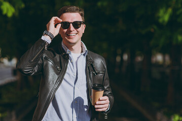 Young smiling stylish man in black leather jacket eyeglasses walk in green park hold takeaway delivery craft paper cup drink coffee to go look camera Concept of people urban lifestyle spring season