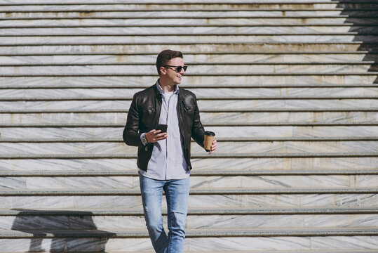 Young Smiling Fun Man Wear Black Leather Jacket Eyeglasses Walk In City Downtown Go Down Concrete Steps Hold Mobile Cell Phone Takeaway Craft Paper Cup Drink Coffee To Go Concept Of Urban Lifestyle