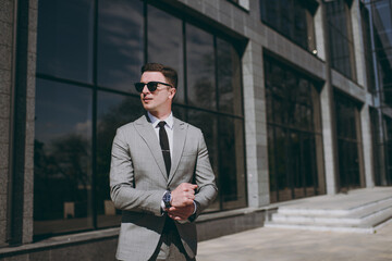 Young satisfied confident european successful employee business man 20s in grey suit walking go near office glass wall building outdoors in downtown city center look aside. Achievement career concept.