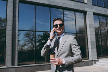 Young happy employee business man 20s in grey suit walk near office glass wall building outdoors in downtown city center hold mobile cell phone takeaway craft paper brown cup coffee to go look camera.