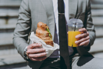 Close up cropped photo portrait shot young caucasian businessman man in grey tie suit sitting on concrete steps in city center eat biting croissant, drink juice have break lunch breakfast outdoors