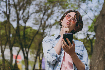 Young dreamful caucasian wistful fun woman 20s wear jeans clothes stand in city spring park outdoors use mobile cell phone chat with friends on social network. People active urban lifestyle concept