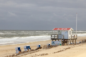 Obraz premium Beautiful beach on the German island of Sylt with white sand, cloudy dark sky, a lifeguard station and beach chairs.