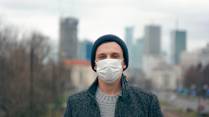 Portrait of Man in Surgical Face Mask Staying Outdside in Big City Downtown and Looking to Camera. COVID-19 Coronavirus Pandemic Outbreak or Smog Background. Close-up picture with Copy Space