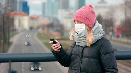 Casual Woman in Surgical Face Mask Uses Mobile App on Smartphone Staying Outdside in Big City Downtown. COVID-19 Coronavirus Pandemic Outbreak or Smog. Picture with Copy Space