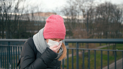 COVID-19 Coronavirus Pandemic Outbreak: Casual Young Sick Woman in Surgical Face Mask and Medical Gloves Sneezes in Street and Coughs covering mouth