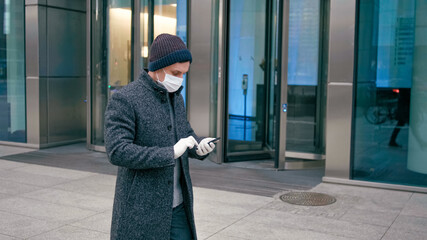 Business Man in Surgical Face Mask Uses Mobile App on Smartphone Walking Outdside in City Downtown Street with Office Buildings. COVID-19 Coronavirus Pandemic Outbreak.
