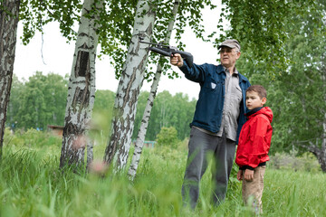A boy in a red windbreaker with a grandfather in a cap who is aiming an air gun in summer park. Concept weapons and children, respect, caution, training.