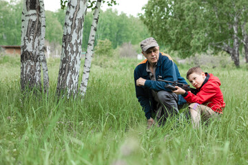 Naklejka na ściany i meble A boy in a red windbreaker is aiming an air gun with a grandfather in a cap in summer park. Concept weapons and children, respect, caution, training.