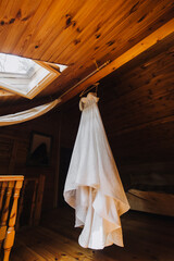 A beautiful, long, white, fashionable dress of the bride hangs on a hanger against the background of a wooden interior in the room. Photography, concept.