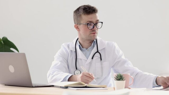 a doctor in a white coat in his office at a medical clinic makes an entry in his diary, a doctor works after seeing patients at a computer, a European man in a doctor's robe makes a record of the