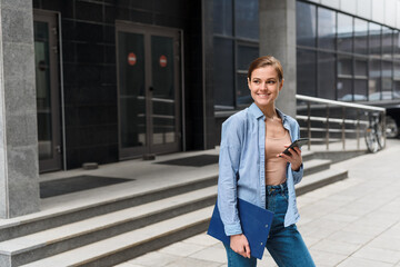 A young stylish businesswoman walks against the background of the business center and holds a folder with documents under her arm, a phone in her hand. Going to work after the coronavirus pandemic