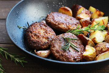 Meat cutlets with fried potatoes and rosemary in a frying pan on a wooden table