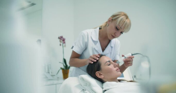 Close-up a blonde female cosmetologist is doing a non-injection bio-revitalization procedure by using a Dermadrop TDA device on the female client who is laying in the cosmetology bed in a bathrobe.
