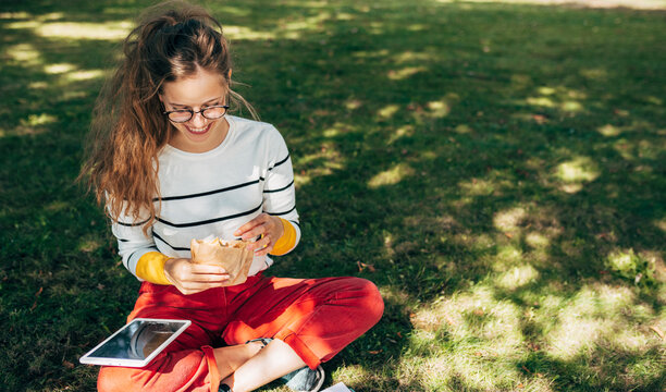 Outdoor horizontal image of a student female sitting on the green grass the college on a sunny day, having a lunch, and studying. Young woman takes a rest eating fast food in the park.