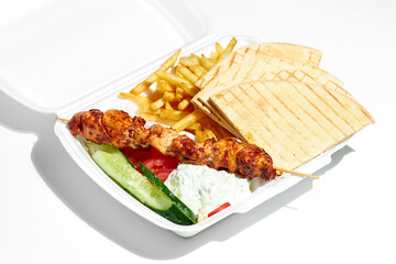 Appetizing Greek souvlaki with cucumbers, fries, pita, yoghurt in a plastic lunch box. Street food, hard light. White background