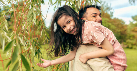 Side view of a cheerful daughter enjoying a ride on the shoulders of her father during the game in the park. Smiling dad playing in the park with his little girl on a sunny day.