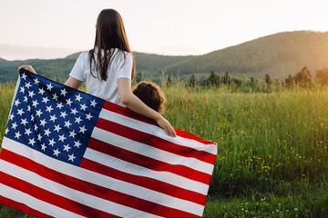 Back view close up of young mother and cute small daughter with national US flag outdoors on background green mountains during sunset. American flag, country, patriotism, independence day 4th july