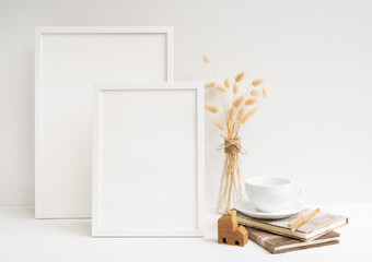 Mock up two poster frames,coffee cup,raft book ,house model and  dried Lagurus ovatus flowers composition in modern glass vase  on  white table and cement wall background