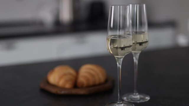 Two Glasses Of Sparkling Wine Sit On The Gray Countertop. On The Background Of The Kitchen. Wooden Bowl With Freshly Baked Croissants.