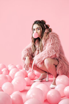 Funny hipster woman blowing bubble with chewing gum sitting in pink balloons background