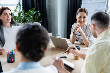 Smiling businesswomen looking at blurred colleague with notebook in office