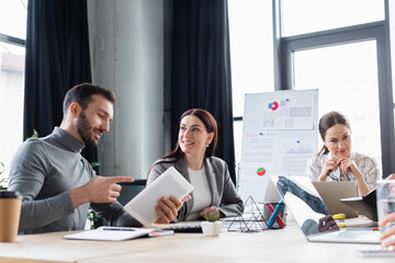 Businessman pointing at digital tablet near smiling colleague at working table in office