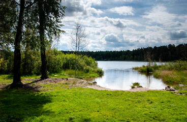 A forest lake, a green lawn in front of a small beach and a dark forest on the far shore.