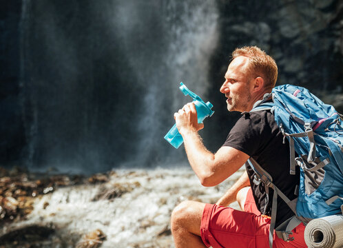 Man with backpack dressed in active trekking clothes drinking the water from touristic flask sitting near mountain river waterfall and enjoying the Nature. Traveling, trekking, or nature concept image